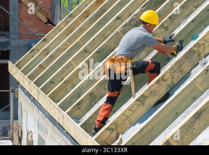 A construction worker with a yellow helmet installs wooden rafters on a building under construction during daylight. Tools are visible in his belt. Stock Photo