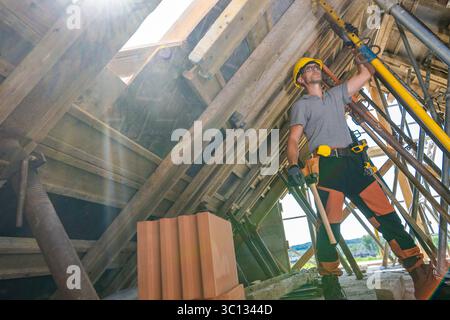 A construction worker uses a lifting device to elevate wooden materials at a sunny job site surrounded by scaffolding. Safety gear is worn for protect Stock Photo