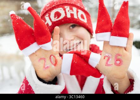 Smiling cheerful child with red Santa hats on fingers and numbers 2026 written on his palms. joy of celebrating New Year and Christmas. Boy waiting fo Stock Photo