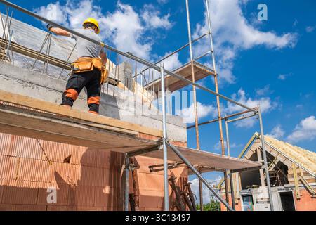 A construction worker works diligently on scaffolding, laying bricks for a new building while surrounded by scaffolding and a clear blue sky overhead. Stock Photo
