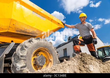 A construction worker in a yellow hard hat stands beside a dump truck at a residential building site, ready to load materials under a clear blue sky. Stock Photo