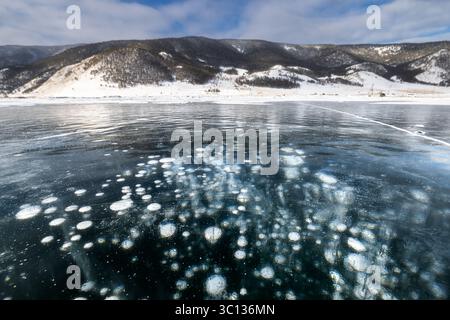 View of frozen methane bubbles trapped beneath the clear, icy surface, with distant, snow-dusted mountains under a bright sky, Baikal, Irkutsk Oblast, Russia. Stock Photo