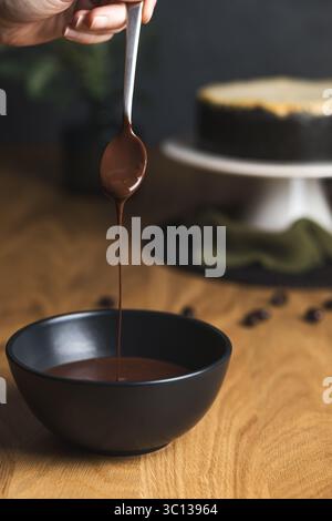 Hand holding a spoon dripping smooth melted chocolate into a black bowl, with a cheesecake on stand in the background. Stock Photo