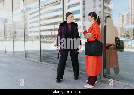 Two businesswomen engaging in a discussion near a modern office building on a bright day Stock Photo