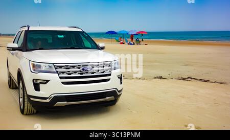 White Ford Explorer SUV parked on a sunny beach with colorful umbrellas and people enjoying a summer day by the calm ocean. Stock Photo