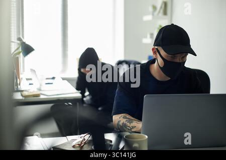 Young Caucasian male hacker wearing cap and mask using laptop at desk in modern office, collaborator in black hoodie breaking into confidential document cabinet to steal data Stock Photo