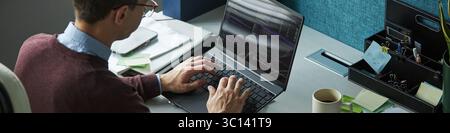 Header of Caucasian young man working on laptop at desk, typing programming code on computer in modern office, wearing glasses, focused on screen, surrounded by stationery Stock Photo