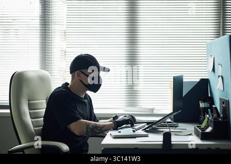Male hacker wearing black mask and gloves sitting at desk typing on computer with tablet and smartphone on table, while stealing confidential business information during break in at company office Stock Photo
