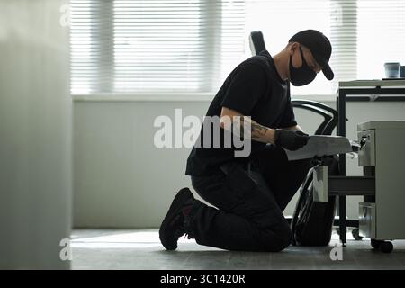 Caucasian young man wearing black mask and gloves kneeling on floor opening drawer and examining documents in modern corporate office, searching for business information Stock Photo