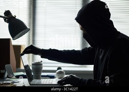 Young male intruder wearing black hoodie, face mask, gloves sitting at desk using laptop, breaking into corporate laptop to access sensitive business information, blinds covering windows Stock Photo