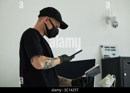 Young Caucasian man wearing black mask and gloves standing in modern office holding smartphone and notebook, checking, scanning sensitive information, tattoos on arms, security camera in background Stock Photo