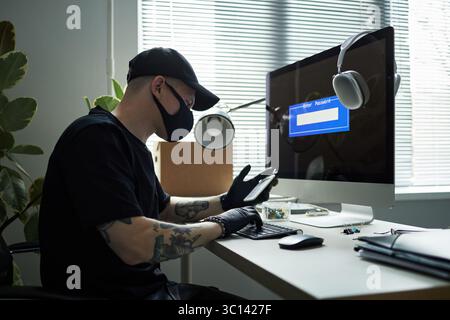 Young Caucasian man wearing cap and black mask sitting at desk using smartphone and typing on keyboard while hacking into computer, with password entry screen visible on monitor in modern office Stock Photo