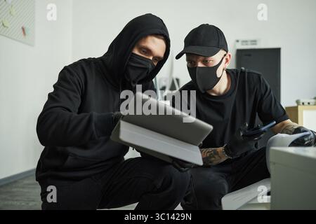 Two male corporate spies wearing black hoodies and face masks examining tablet device together indoors, one holding smartphone, both crouching and focusing on screen, while raiding company office Stock Photo