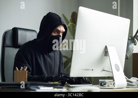 Middle aged man wearing black hoodie, gloves and mask sitting at desk, hacking into corporate network using desktop computer, while committing cybercrime and data theft Stock Photo