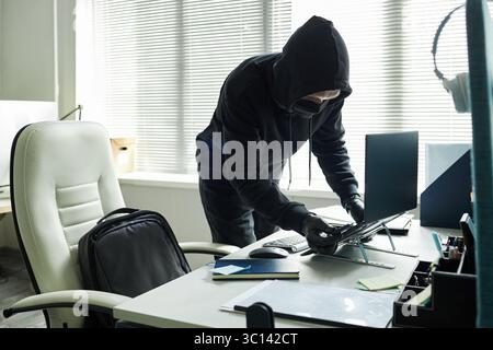 Anonymous man wearing hood. black mask and gloves stealing laptop from modern office desk, standing next to open backpack and looking down at device, blinds covering window in background Stock Photo