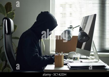 Young man wearing black hooded sweatshirt, obscuring face, sitting at desk, hacking into desktop computer in corporate network while breaking into company office, typing commands on keyboard Stock Photo