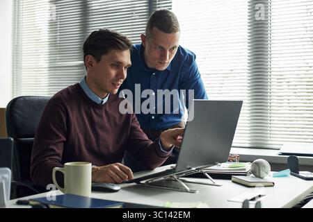 Caucasian young men collaborating at desk, one sitting and pointing at laptop screen while other standing and observing, working together in modern office environment Stock Photo