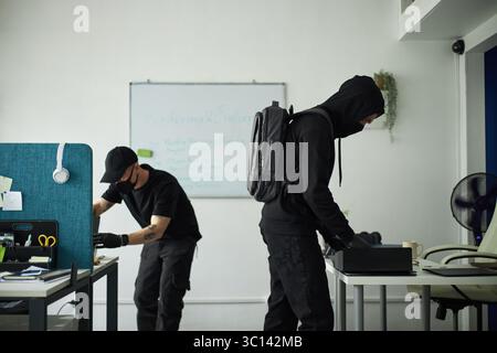 Two young male spies wearing black clothing and gloves, searching through office desks and drawers, stealing corporate electronic gadgets, searching for confidential documents Stock Photo