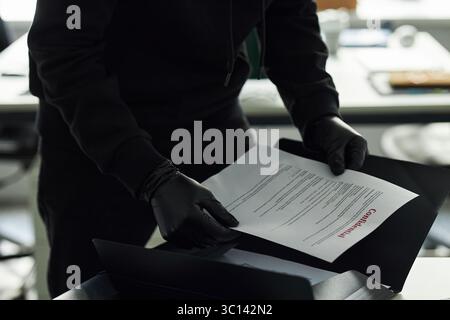 Unrecognizable man wearing black clothes and gloves handling confidential document in office setting, searching for sensitive business information in folder, only upper body and hands visible Stock Photo