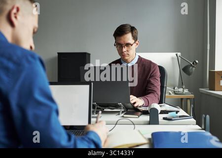 Caucasian young male manager working on laptop at desk in modern office, sitting across from colleague taking notes, both focused on tasks, technology visible on desks Stock Photo