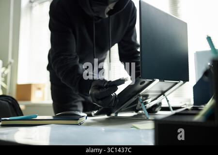 Unrecognizable male intruder wearing black gloves stealing data by inserting USB flash drive with malware into laptop computer on office desk to steal data, face obscured by hood Stock Photo