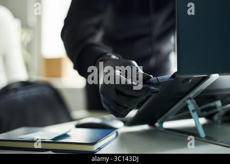 Anonymous man wearing black gloves inserting USB flash drive into laptop computer, performing data transfer, theft, malware or cyber fraud in modern corporate office Stock Photo