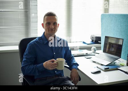 Portrait of young Caucasian man sitting at desk holding coffee mug, looking into camera, working in modern office with open laptop, documents and office supplies visible Stock Photo