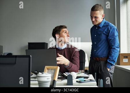 Caucasian young adult man sitting at desk explaining project to team member standing nearby in modern office workspace with laptops and documents visible, collaborating on presentation or report Stock Photo