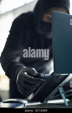 Unrecognizable man wearing black gloves and hoodie inserting USB flash drive with malware into laptop to hack corporate computer system, engaging in covert criminal activity at workspace Stock Photo