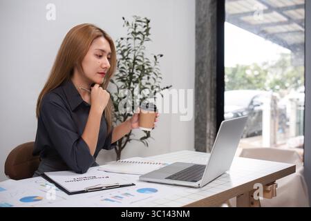 Young Professional Woman Analyzing Work Data in Modern Office Setting with Laptop and Coffee Stock Photo