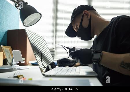 Male hacker wearing black cap and face mask sitting at desk in office, breaking into corporate laptop to steal information from computer system using scanning tools, holding cable in gloved hands Stock Photo