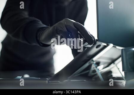 Unrecognizable man wearing black gloves breaking in and accessing laptop in office, inserting flash drive to steal data, concept of online fraud, cybersecurity or identity theft Stock Photo