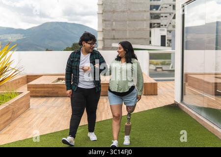A young man helps a disabled woman with a prosthetic leg as they walk together through a rooftop garden. They are married, and she is pregnant. Stock Photo