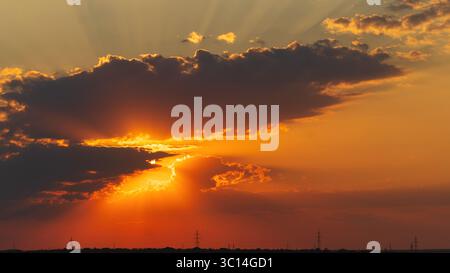 Golden rays from a setting sun shine past the edges of dark clouds, illuminating the sky above a silhouetted horizon dotted with power lines Stock Photo