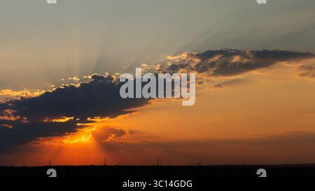 A wide panorama shows a sunset with glowing sun rays shining from behind a dark cloud, silhouetted horizon, and faint power lines under a colorful sky Stock Photo