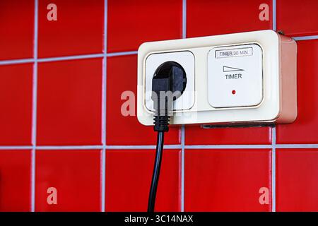 A closeup of a black electrical plug inserted into a beige timer switch outlet mounted on a red tiled wall. Stock Photo