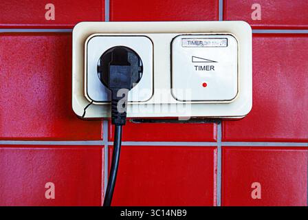 Closeup of a cracked beige power socket with a timer switch and plugged in black cable, mounted on a red tiled kitchen wall. Stock Photo