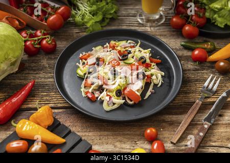 Fresh greek salad with tomato, cabbage, olive and cheese in the black ceramic plate near one glass orange juise on the rustic wooden table, brown back Stock Photo