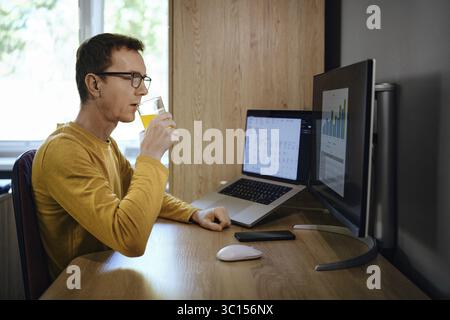 Individual is focused on work at a home office, sipping a drink while looking at two computer screens displaying charts. The workspace is organized wi Stock Photo