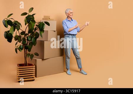 Mature woman standing in casual attire next to stacked cardboard boxes, gesturing energetically on a beige background with a plant Stock Photo