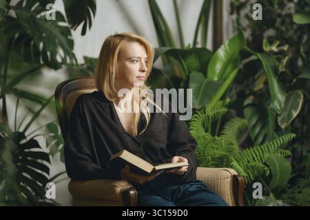 A woman sits comfortably in an armchair holding a book, surrounded by vibrant green plants.She is distracted from her reading and is looking thoughtfu Stock Photo