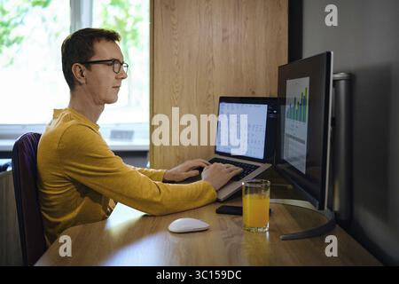 A middle-aged man focused on his work sits at a wooden desk with two computer screens. He enters various data and looks at the chart, analyzing and pr Stock Photo
