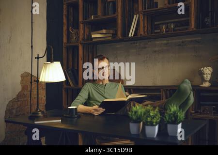 A middle-aged man relaxes with his feet up on a desk in a home office, reading a book. Natural light illuminates the cozy space, filled with books and Stock Photo