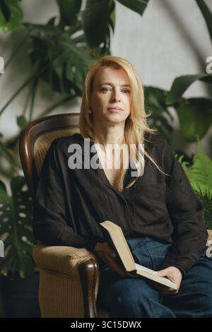A woman with long blonde hair sits comfortably in a vintage armchair holding a book. She is surrounded by vibrant greenery, creating a serene and invi Stock Photo