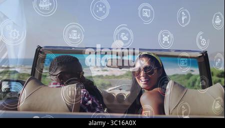 Smiling couple turning heads toward camera in cream convertible on coastal road, tech icons Stock Photo
