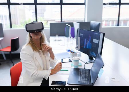 VR headset resting on office desk with dual monitors showing graphs and coffee cup near window Stock Photo