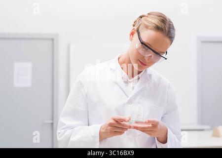Female lab technician examining glass slide in sterile lab wearing lab coat and goggles Stock Photo