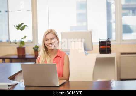 Female worker sitting at wooden desk in bright office, typing on silver laptop near file organizer Stock Photo