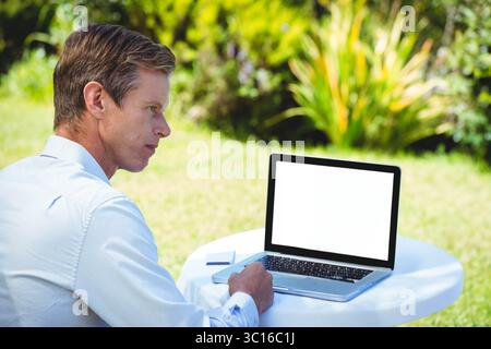 Laptop sitting on round plastic table on lawn near garden shrubs, with notepad, pen and smartphone Stock Photo