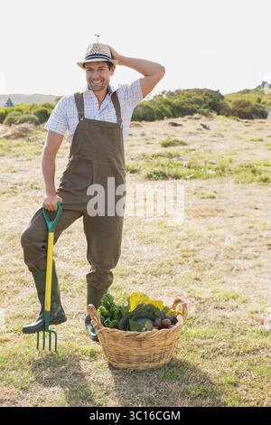 Man standing behind wicker basket holding pitchfork in dry field harvesting seasonal vegetables Stock Photo